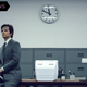 Adam Scott sits stiffly on a desk, wearing a grey suit and a blue bandage on his forehead, in a scene from Severance.